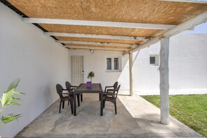 Une terrasse extérieure avec une table et des chaises, une pergola en bois, une plante en pot et un bâtiment blanc en arrière-plan.