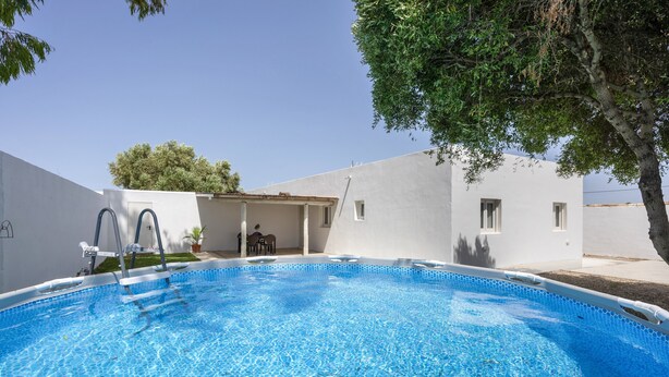 A swimming pool with a clear blue water surface, surrounded by a white building with a flat roof and a tree to the right.