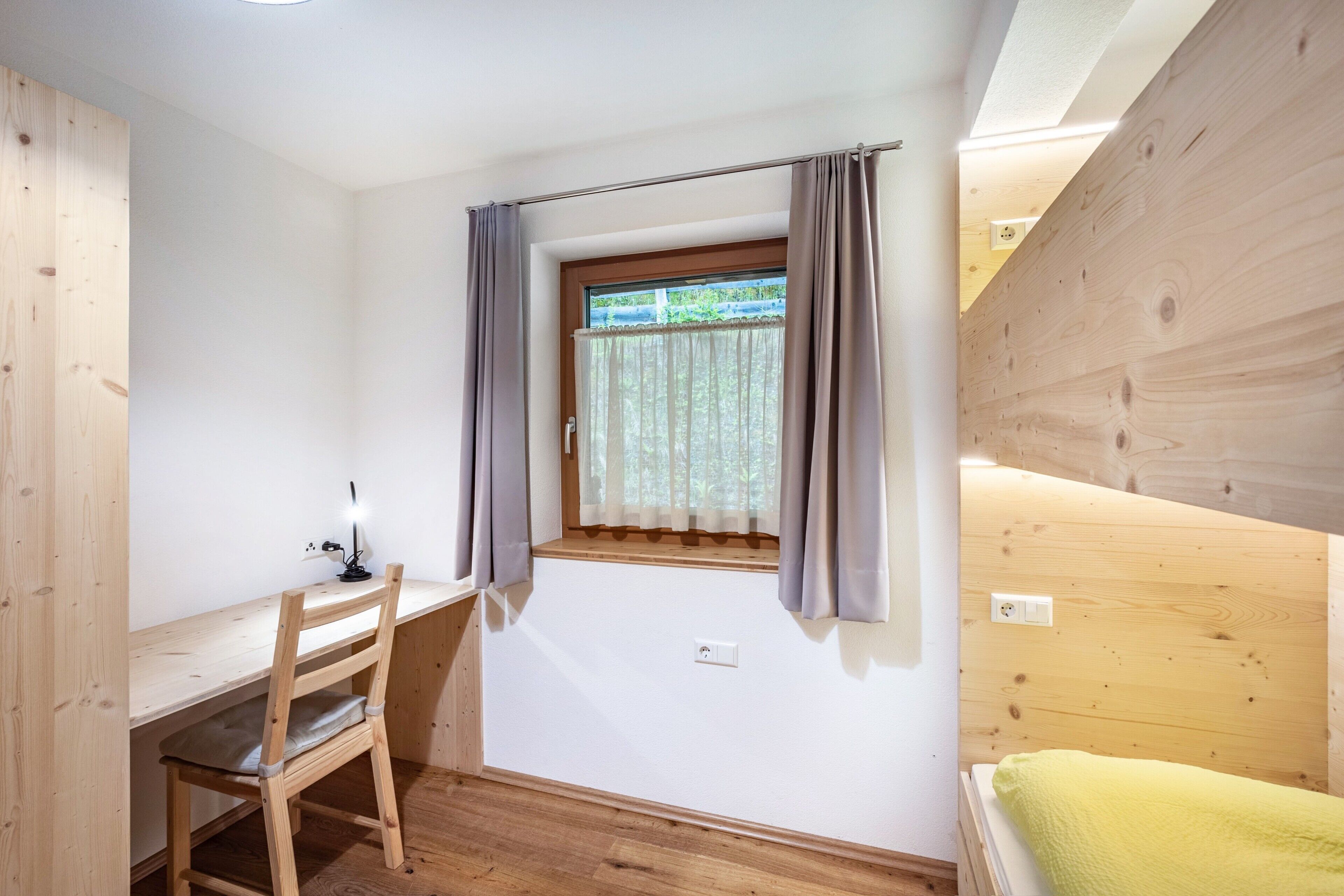 Une pièce avec un bureau en bois, une chaise, une fenêtre avec des rideaux et un lit recouvert d’une couverture verte.