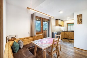 A cozy room with a wooden bench, a table with a plastic cover, a kitchen area with wooden cabinets, and a window with a view of mountains.