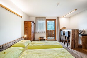 A bedroom with a bed, a wooden headboard, a desk with chairs, a small stool, and a view through a window.
