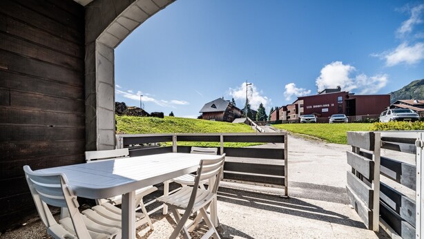 Een terras met een tafel en stoelen, een houten gebouw op de achtergrond en een heldere lucht.