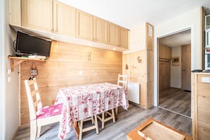 Una habitación con paneles de madera, una mesa de comedor, sillas, un televisor en un estante y una pequeña cocina.