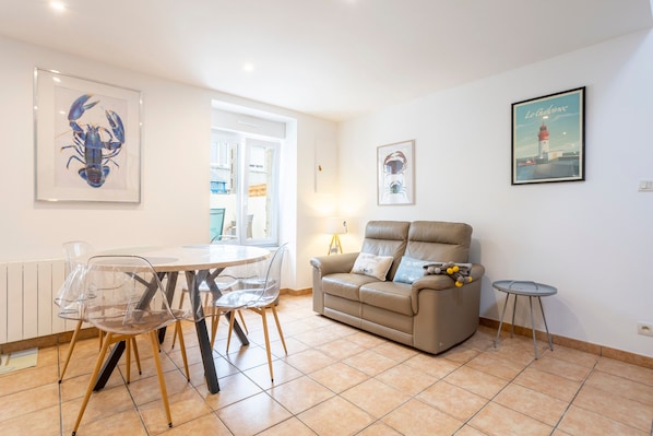 Un salon avec un canapé beige, une table à manger entourée de chaises transparentes et des œuvres d’art accrochées aux murs.