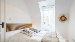 Un dormitorio con cabecera de listones, cama con ropa blanca, ventana con cortinas y un adorno de nido colgado en la pared.