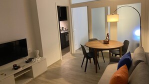 Un salon moderne avec un téléviseur à écran plat, une table à manger avec des chaises, un canapé avec des coussins, un lustre et, au fond, une cuisine.