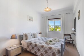 Quarto com cama, mesas de cabeceira, cadeira, janela com cortinas e luminária pendente.