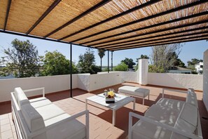 Un espacio de estar al aire libre cubierto con mobiliario blanco, una mesa con bebida y fruta, y vistas del paisaje circundante.