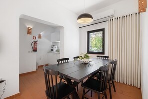 Un comedor con una mesa de madera oscura y sillas negras, una ventana con cortinas y una cocina al fondo.