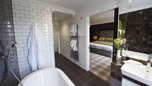 A modern bathroom with a bathtub, a shower, and a large mirror. There is a bed visible in the background, suggesting the bathroom is part of a hotel room. - Cbh Ramside Hotel Golf Spa Durham (Durham)