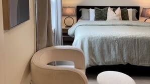 Un dormitorio con cama, una silla, un pequeño reposapiés, un cuadro y una ventana con cortinas.