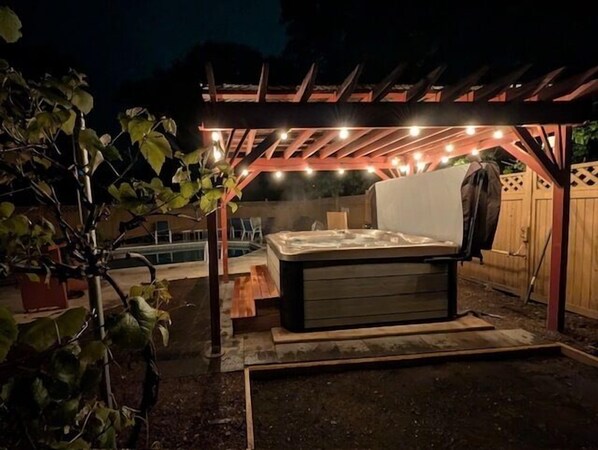 A hot tub under a wooden pergola with string lights, surrounded by a garden and a pool.