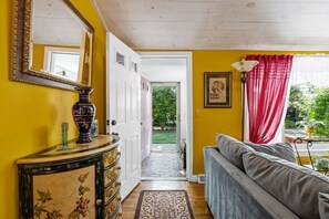 A room with a yellow wall, a large mirror, a dresser with floral patterns, a gray sofa, and a red curtain. - Beach Pass Included! (Barnstable)