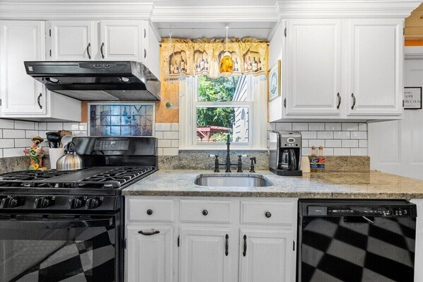 A kitchen with white cabinets, a black stove, a sink, and a window with a view of greenery. - Beach Pass Included! (Barnstable)