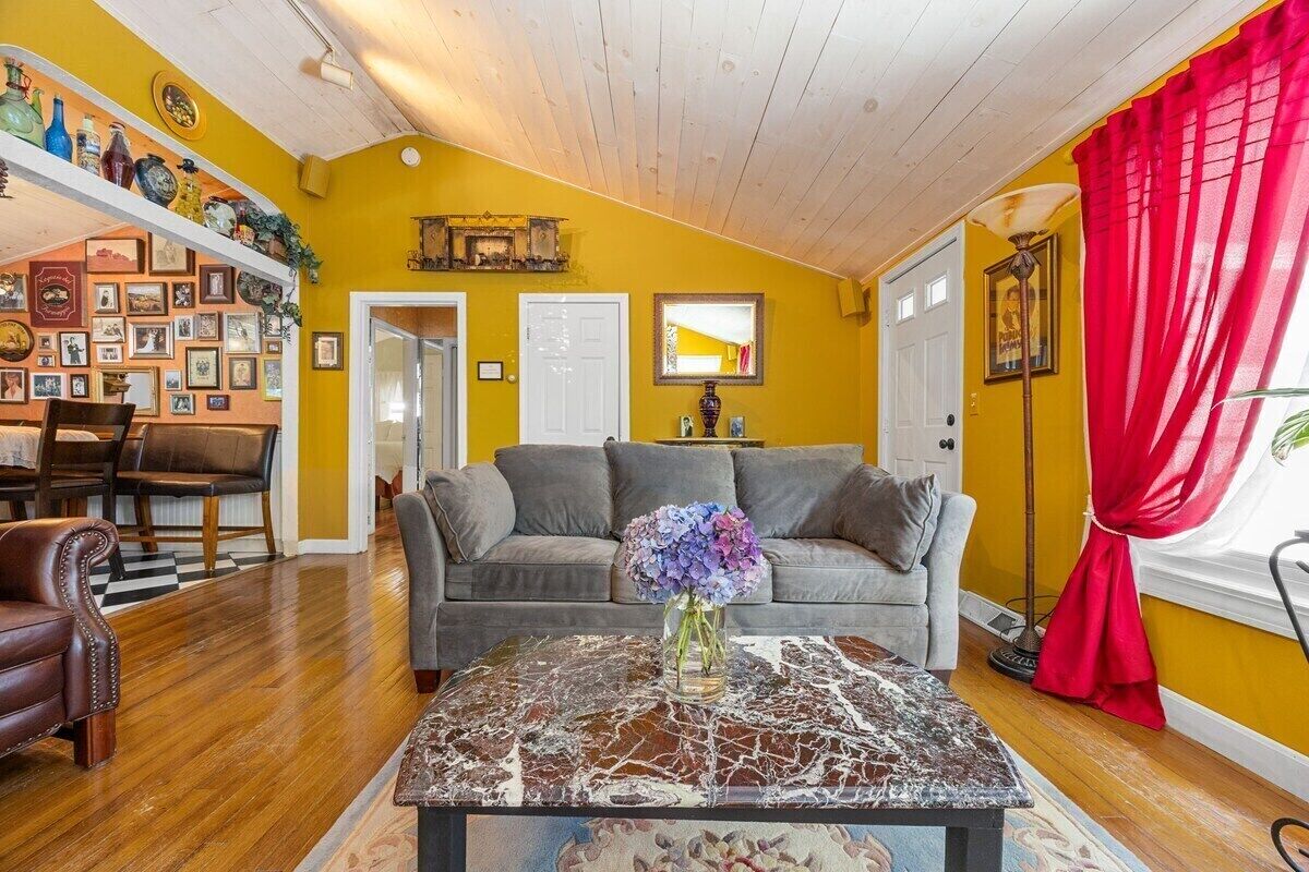 A living room with a sofa, a coffee table with a vase of flowers, a dining area with chairs, and a wall decorated with pictures and a shelf with bottles.