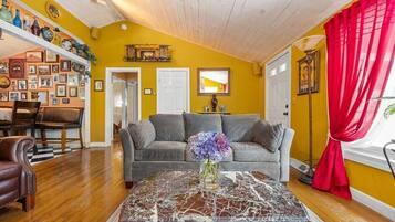 A living room with a sofa, a coffee table with a vase of flowers, a dining area with chairs, and a wall decorated with pictures and a shelf with bottles.