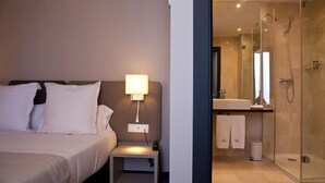 Ett hotellrum med en säng, ett nattduksbord, en lampa och en duschavdelning med glasväggar, handfat och handduk.