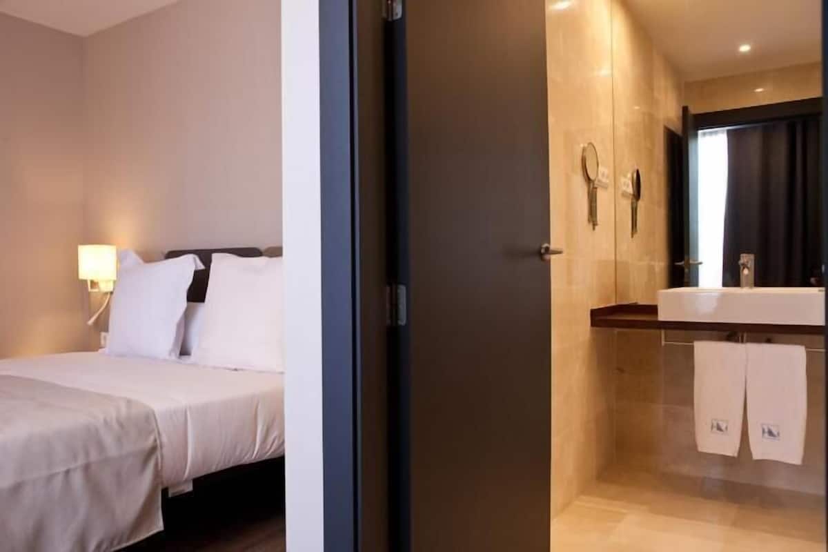 A hotel room with a bed, a bedside lamp, and a bathroom with a sink and mirror.