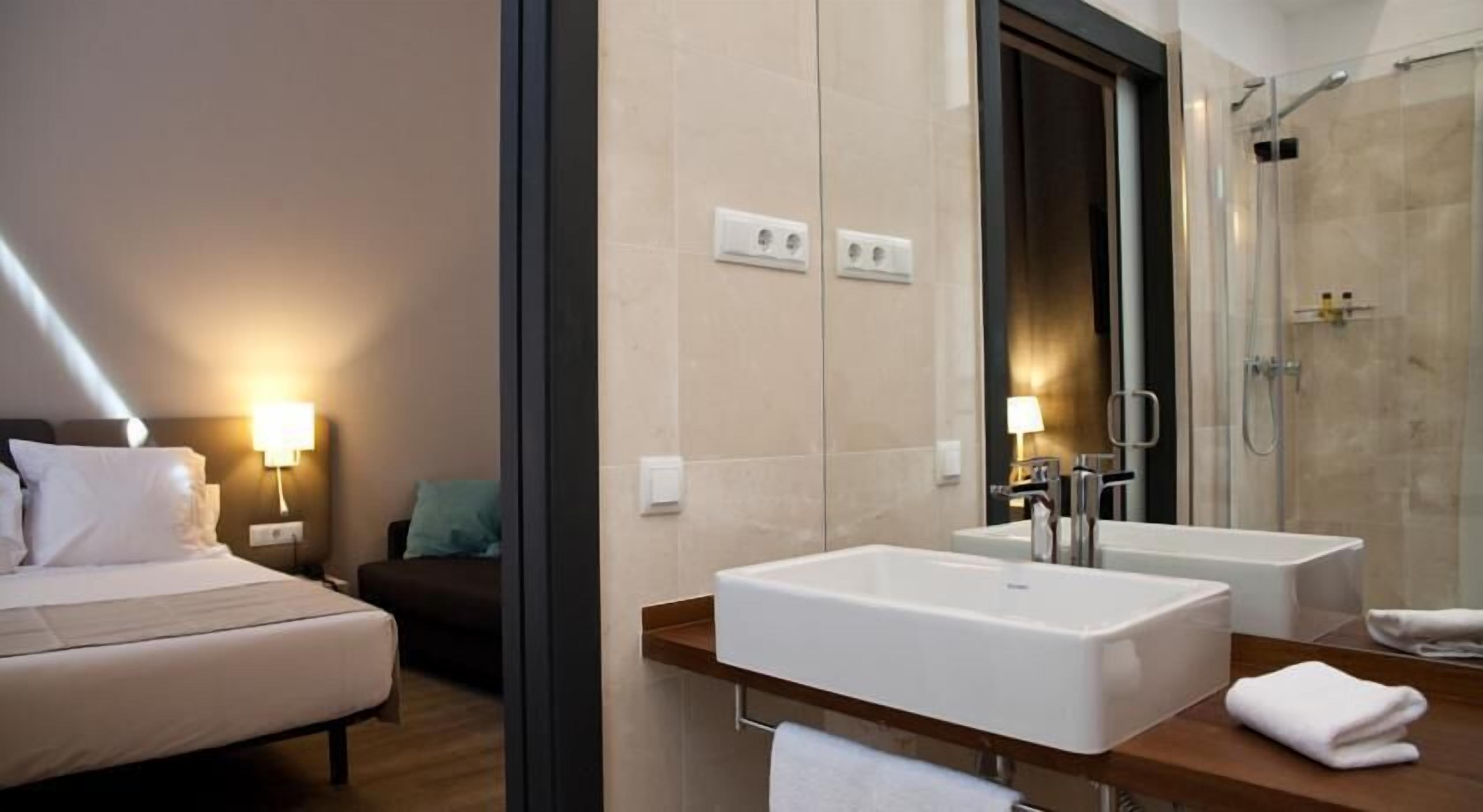 A hotel room with a bed, bedside lamp, a small table with a chair, a large mirror, two sinks, and a shower area.