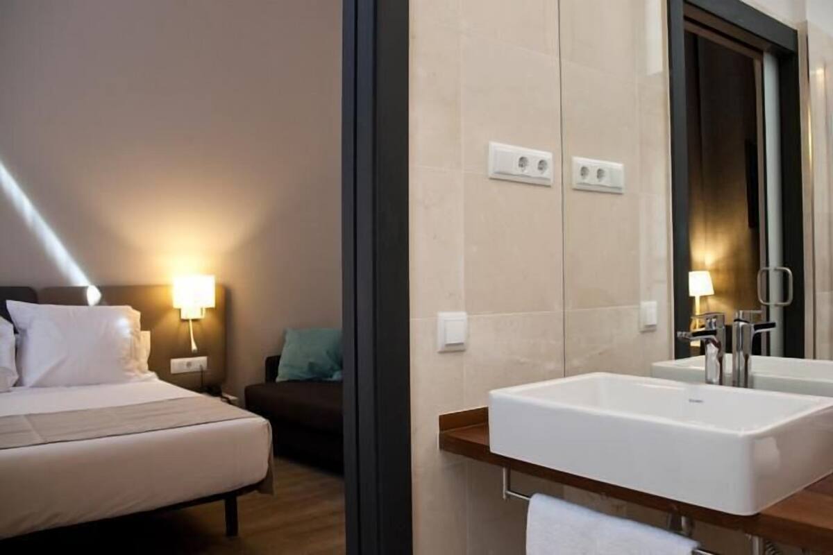 A hotel room with a bed, bedside table, lamp, and a bathroom with a shower, two sinks, and a mirror.