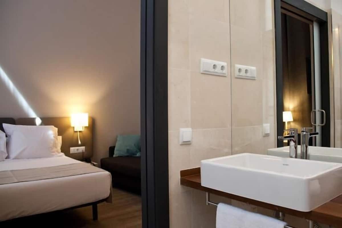 A hotel room with a bed, bedside lamp, a small table with a chair, a large mirror, two sinks, and a shower area.