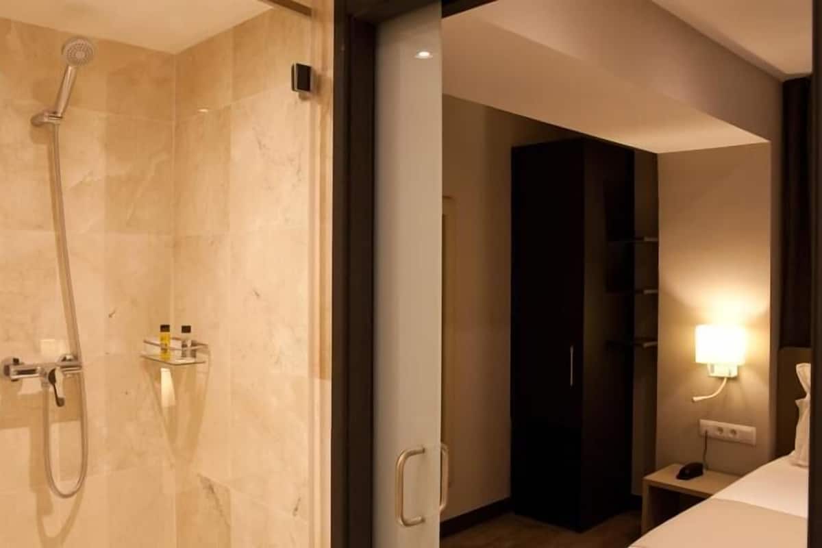 A modern bathroom with a glass-enclosed shower, a vanity with a mirror, and a bedside table with a lamp.