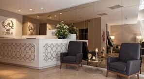 A reception area with a modern design, featuring a reception desk with a logo, two armchairs, a small table with candles, and a large floral arrangement.