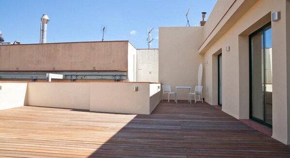 A rooftop terrace with a wooden deck, two white chairs, and a glass door leading to an interior space.