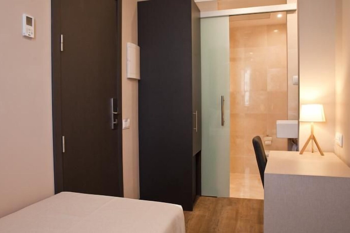 Habitación de hotel con cama, escritorio y silla. Se observa una puerta que conduce a otra habitación y una lámpara fijada a la pared.