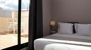 Habitación de hotel con cama, mesita de noche, lámpara y vistas a un balcón exterior con mesa y sillas.