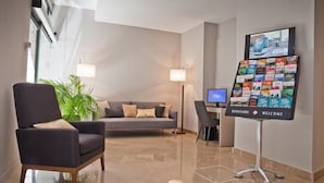 Una hall d'albergo moderna con un divano grigio, una poltrona scura, una scrivania con un computer e un bancone accoglienza con diverse card.
