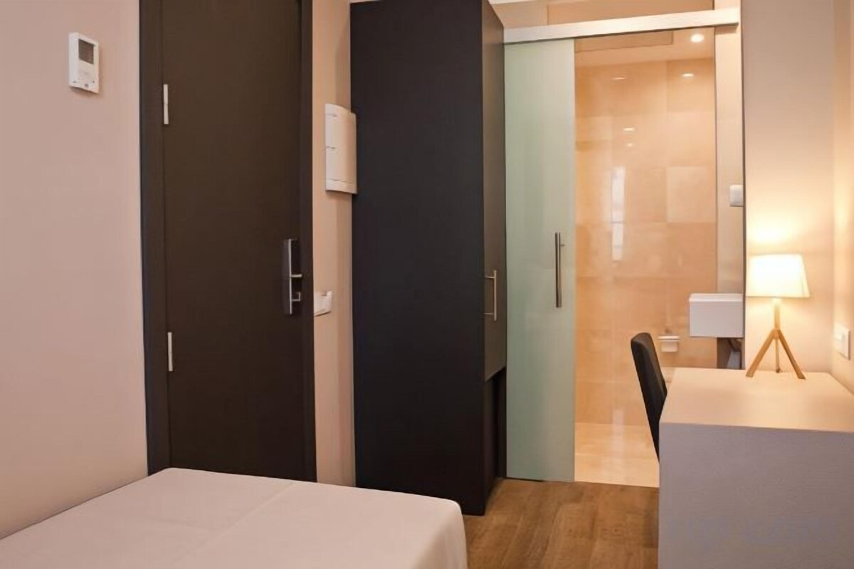 A hotel room with a bed, a desk, and a chair. There is a door leading to another room.