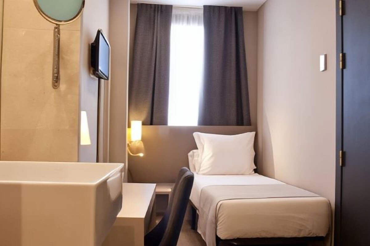 A compact hotel room with a single bed, a small desk, a mirror, and a window with curtains.