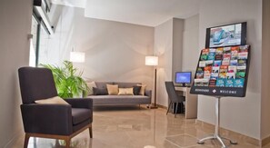 En moderne hotellobby med en grå sofa, en mørk lænestol, et skrivebord med en computer og en velkomststander med forskellige kort.