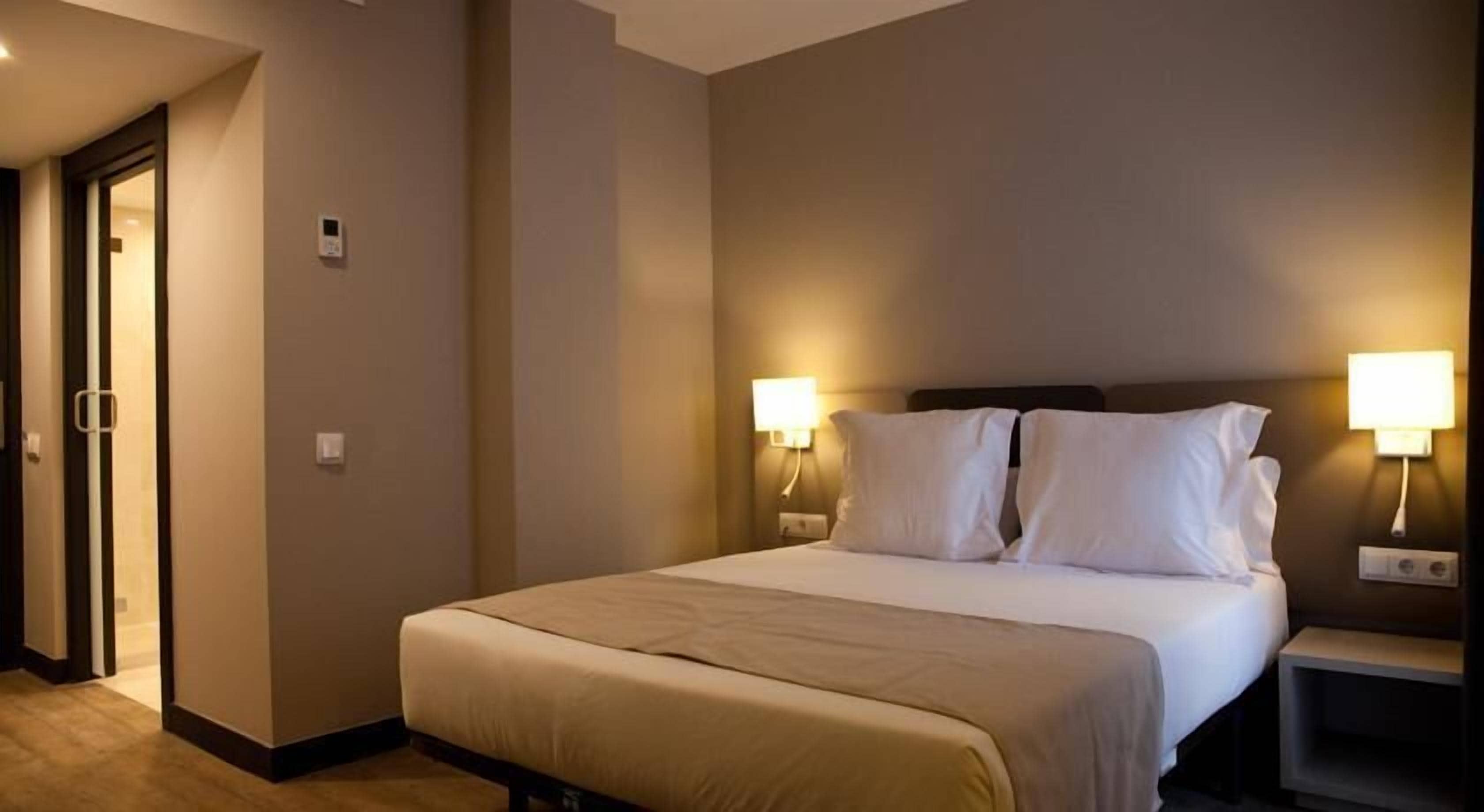 A hotel room with a bed, two bedside lamps, and a door.