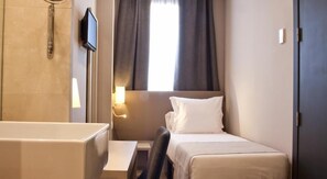 Une chambre d’hôtel avec un lit simple, un petit bureau, une chaise, une salle de bain avec baignoire et un téléviseur fixé au mur.