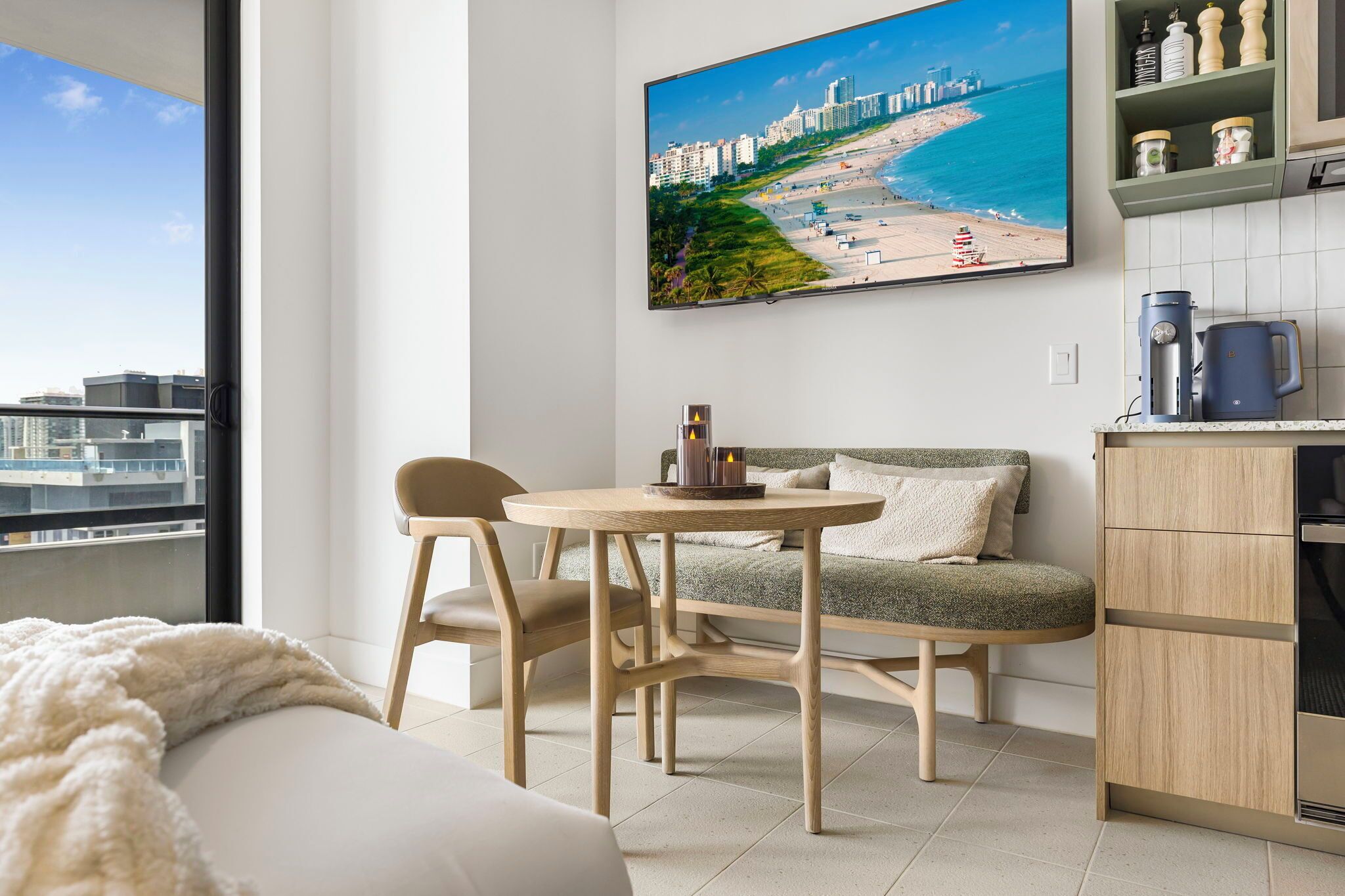 A modern living room with a sofa, a round table, and chairs. A large window offers a view of a cityscape and beach.
