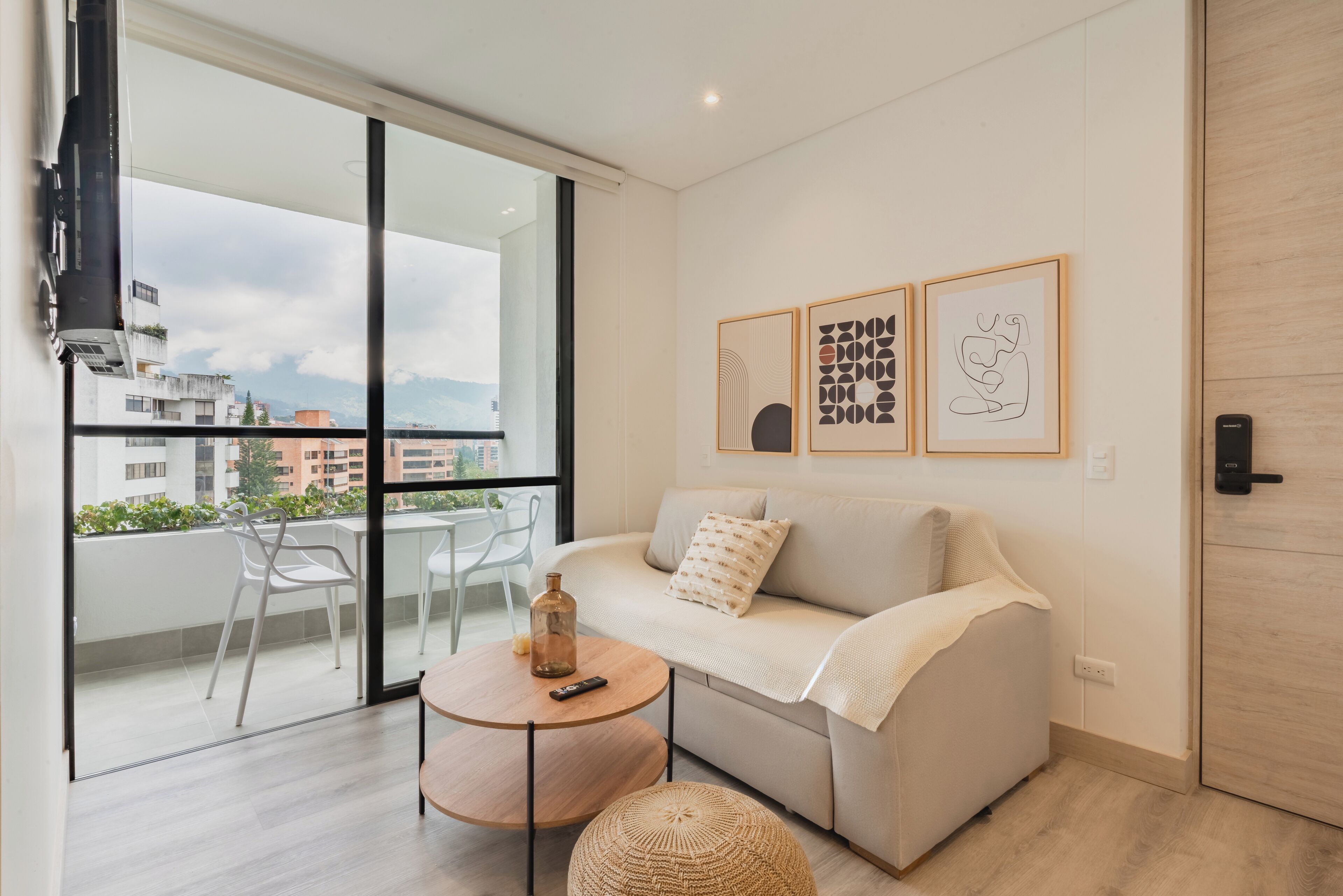 A modern living room with a sofa, a round coffee table, and a balcony with chairs. There are framed artworks on the wall and a door with a lock.