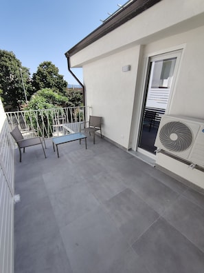 A balcony with a table and chairs, a white wall, and a window.