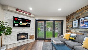 Una sala de estar moderna con un televisor de pantalla plana mostrando servicios de streaming, una chimenea, un sofá gris con cojines amarillos y una pared de madera.