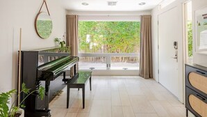 A spacious room with a piano, a bench, and a large window with a view of greenery. - Charming 2-bedroom Mid-Century Modern house in enchanting Palm Springs (Palm Springs)