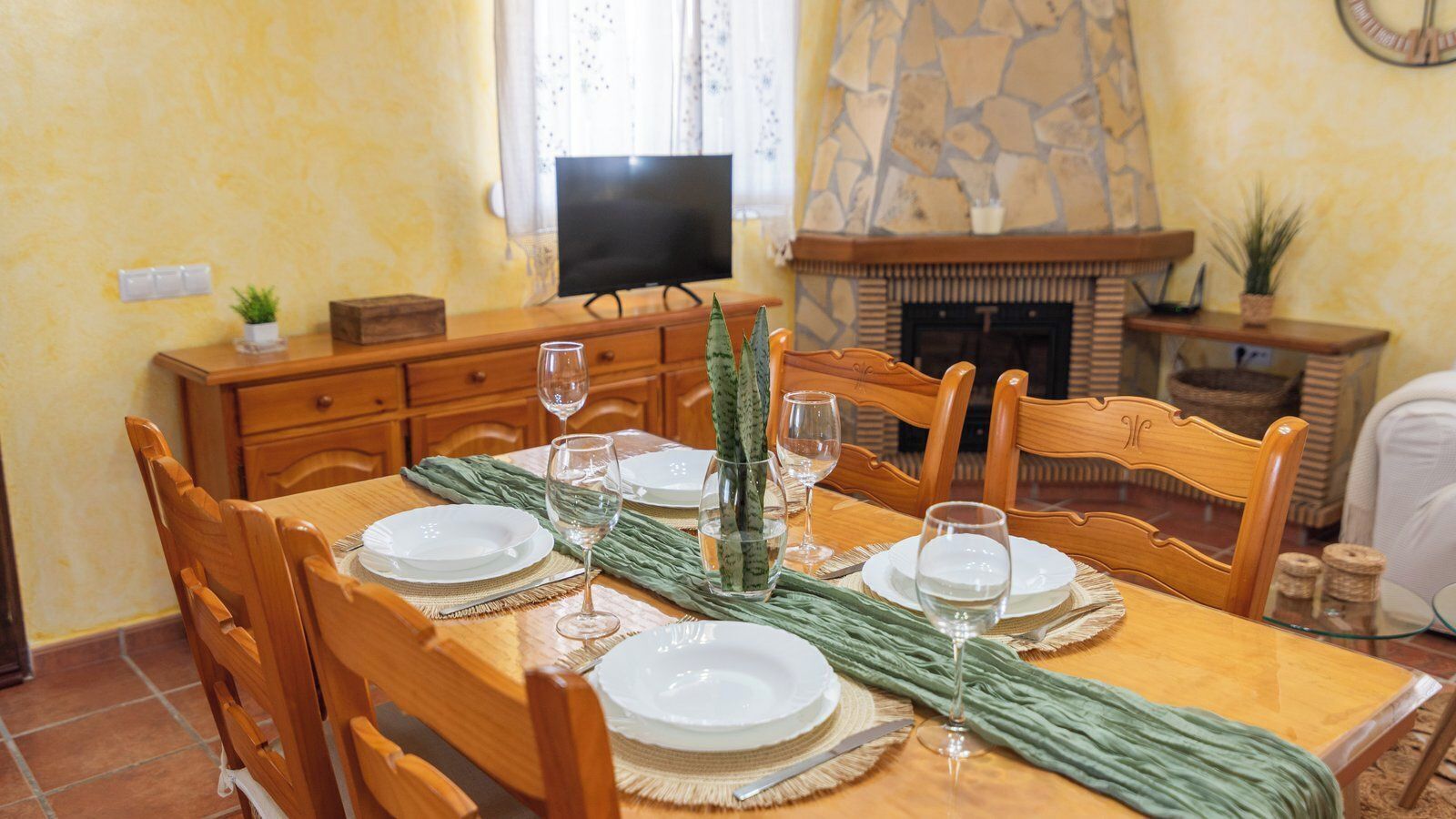 Una mesa de comedor dispuesta con platos, vasos y un centro de mesa. Se ve un televisor y una chimenea al fondo.