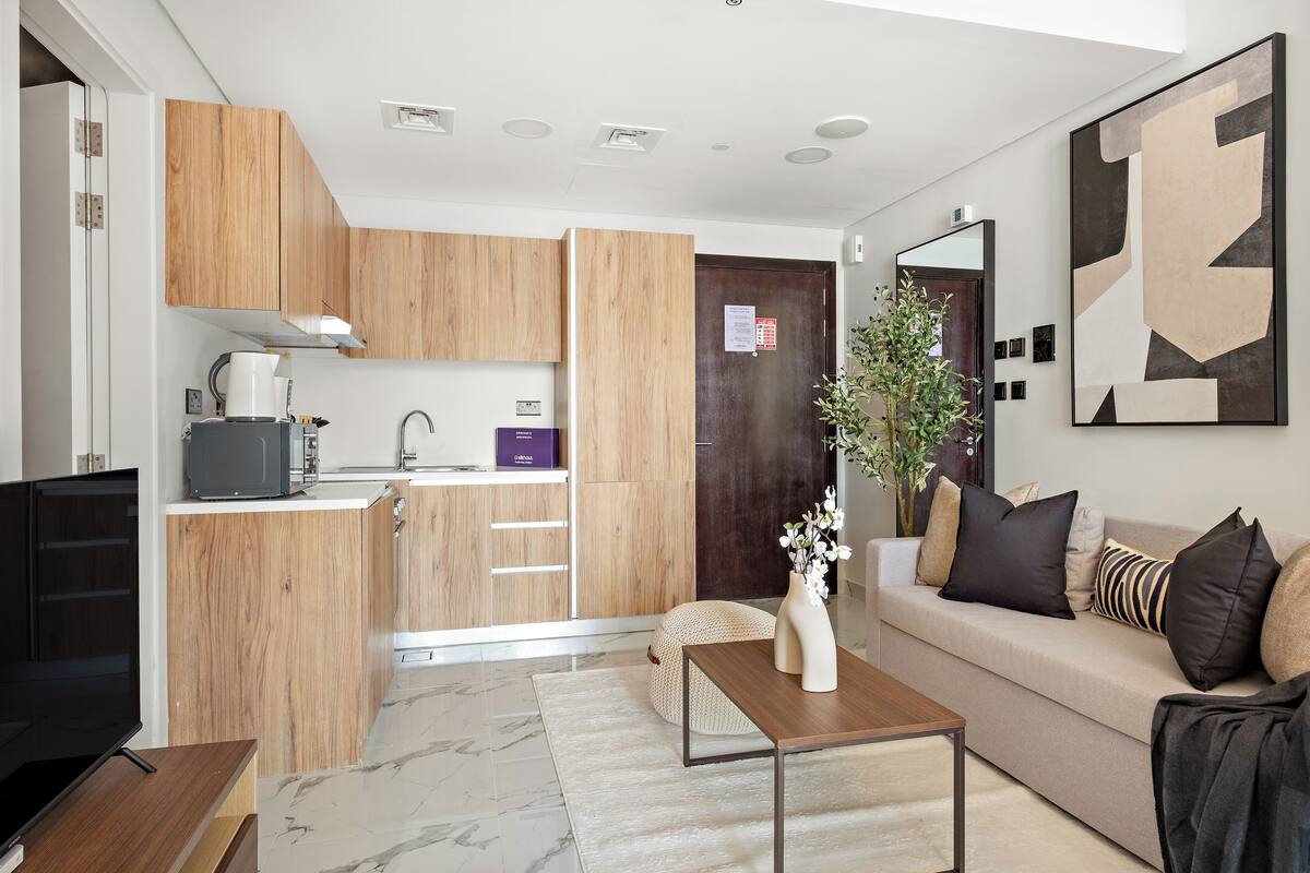 A modern living room with a sofa, coffee table, and a TV. There is a kitchen area with wooden cabinets and a microwave.