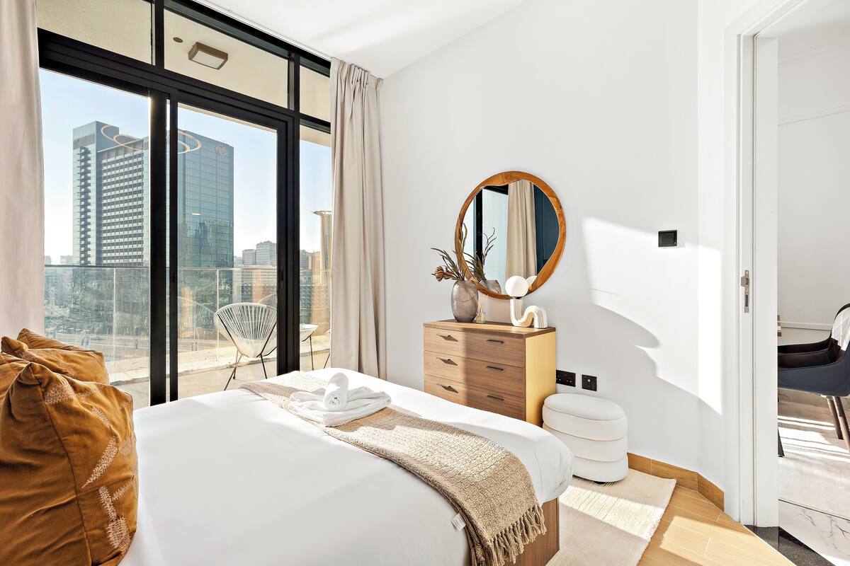 A modern hotel room with a large bed, a wooden dresser, a round mirror, and a view of the cityscape through large windows.