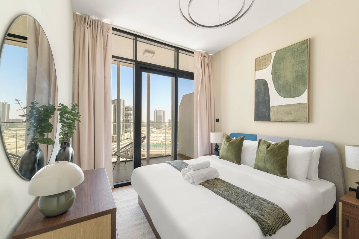 A hotel room with a large bed, a view of the city, a modern lamp, and a large mirror.