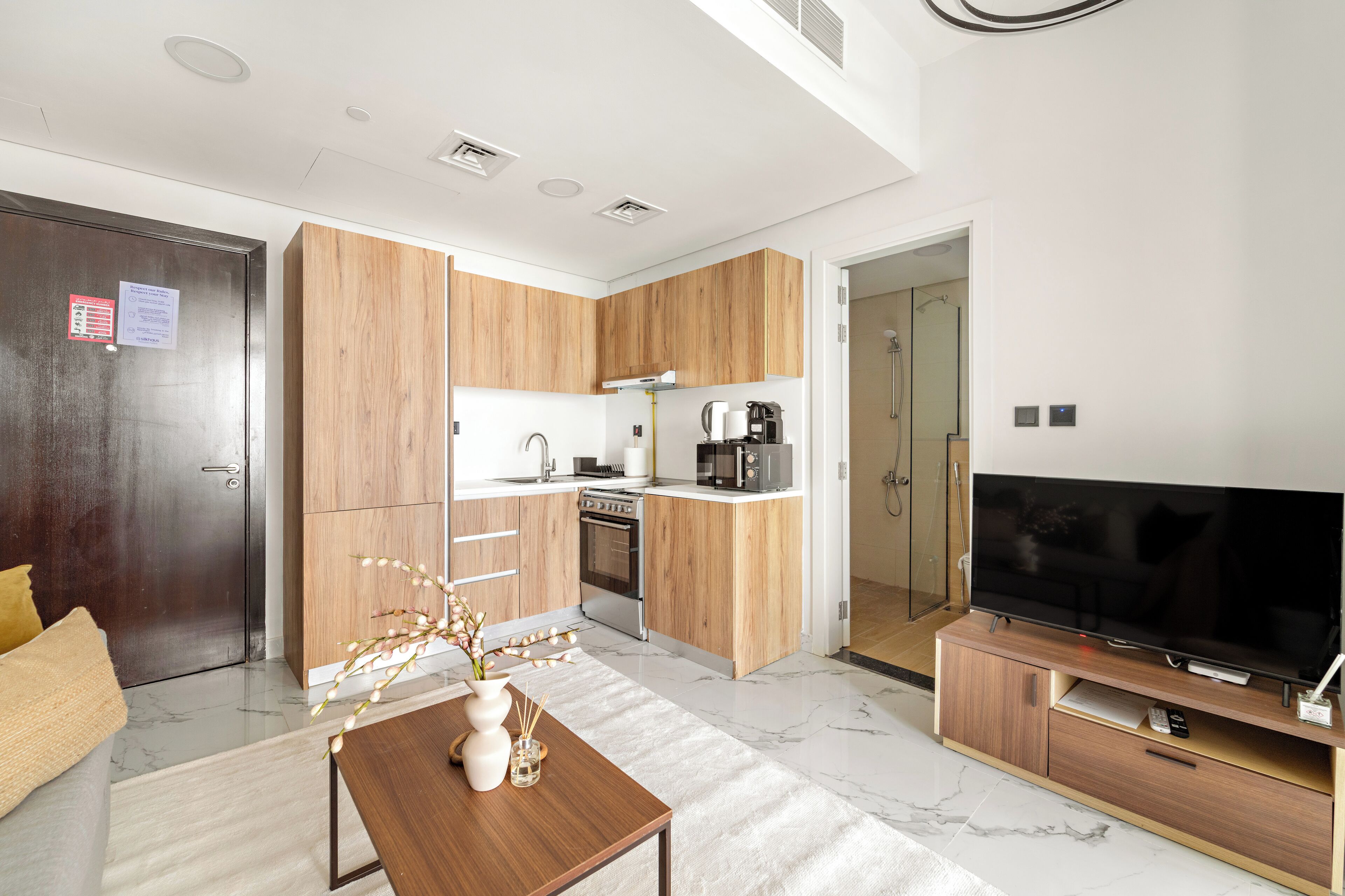 A modern kitchen with wooden cabinets, a built-in oven, and a microwave. There is a dining area with a table and chairs, and a large flat-screen TV on a wooden stand.