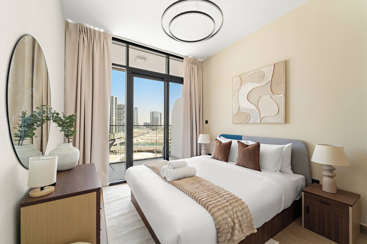 A modern hotel room with a large bed, a desk, and a view of the cityscape through floor-to-ceiling windows.