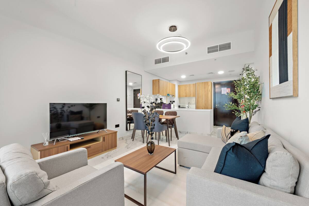 A modern living room with a flat-screen TV, a wooden coffee table, and a sofa. There is a dining area with a table and chairs in the background.