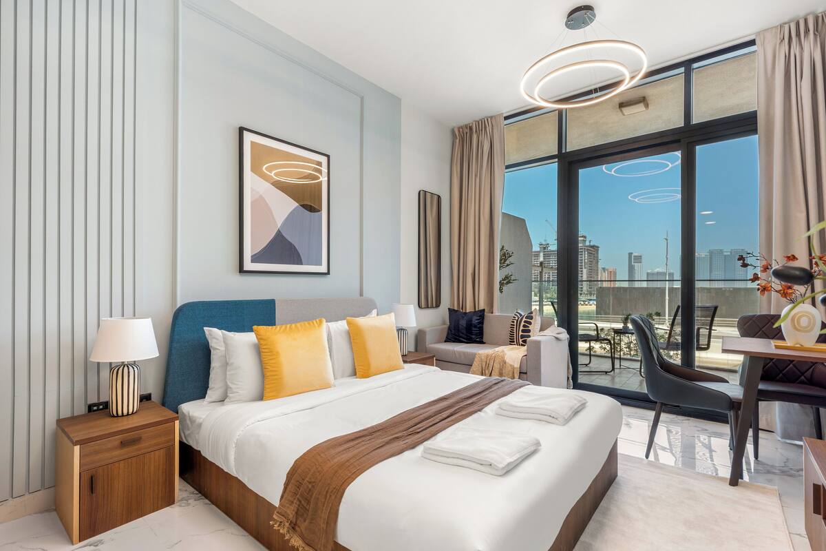 A modern hotel room with a large bed, a desk, and a view of the city through large windows.