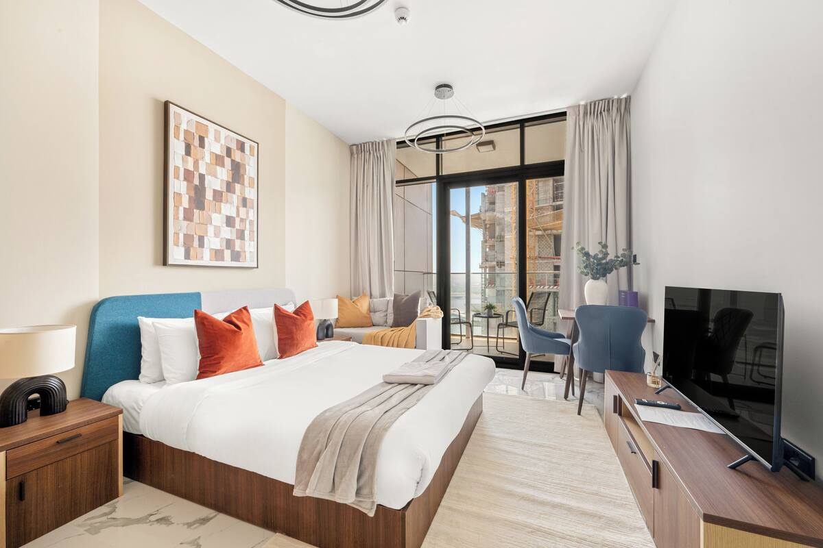 A modern hotel room with a large bed, a desk with a flat-screen TV, a chair, and a balcony with a view of the city.